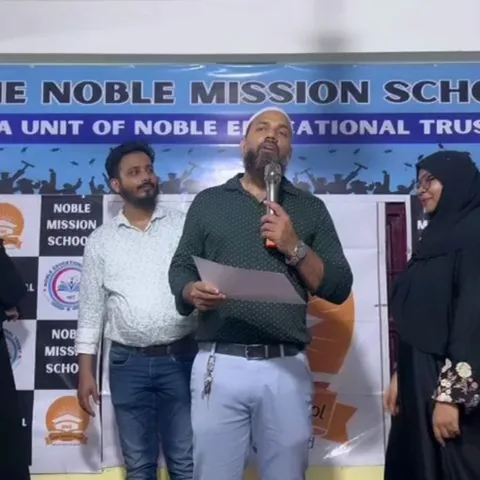 Speaker with a microphone on stage in front of a The Noble Mission School banner at prize distribution